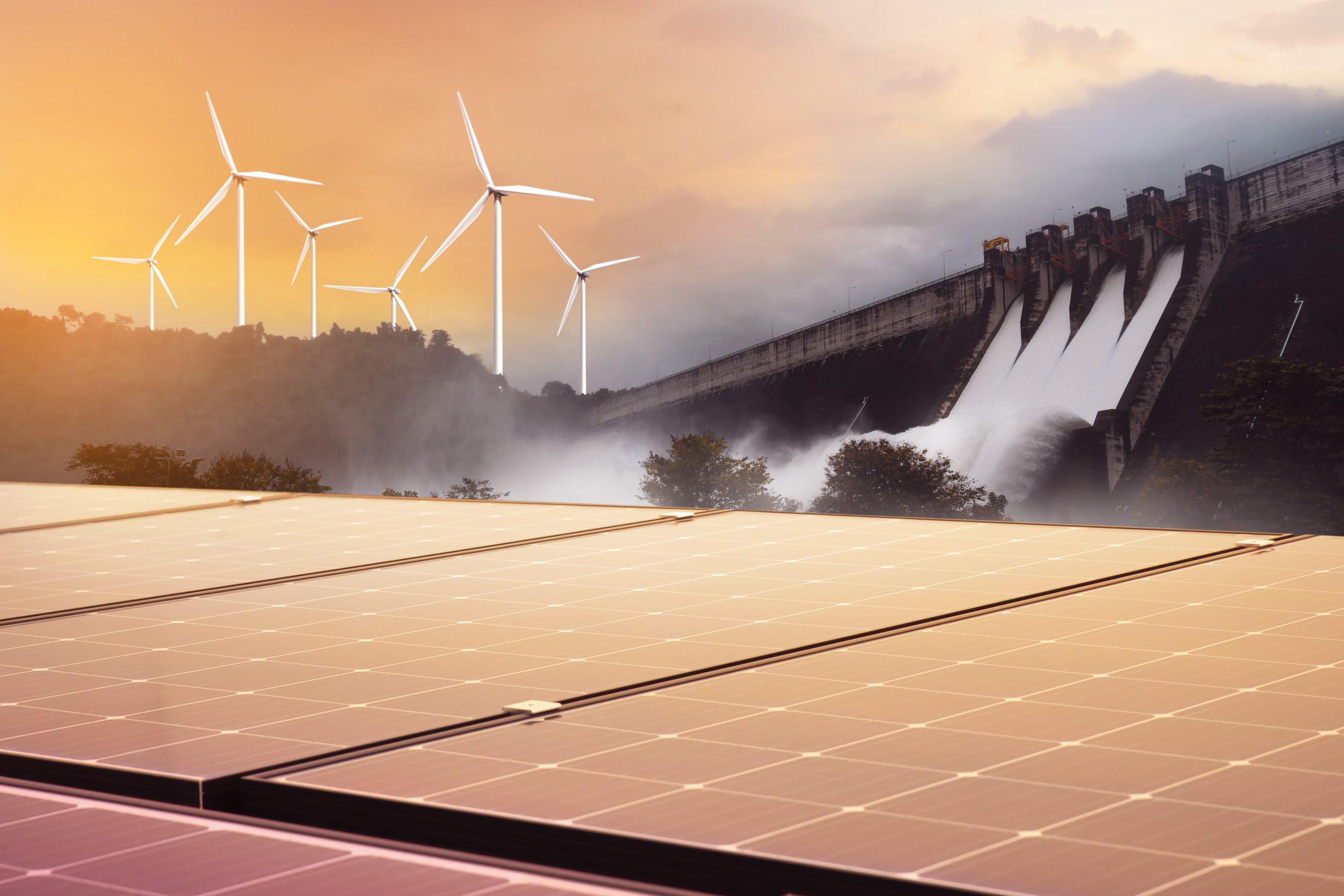 Solar panels in the foreground with wind turbines and a hydroelectric dam behind them at sunset.