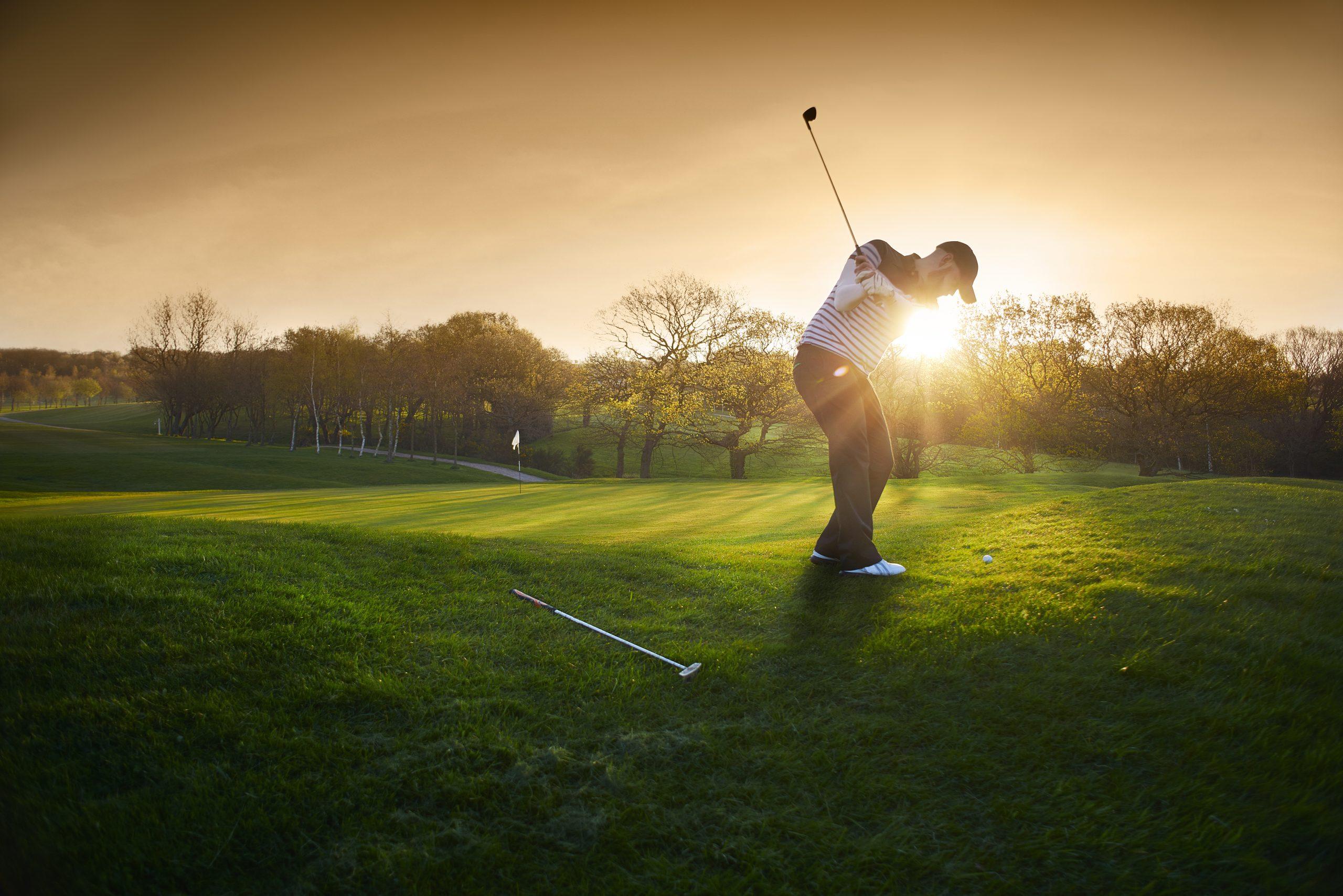 A golfer at the top of their backswing on a fairway at sunset.